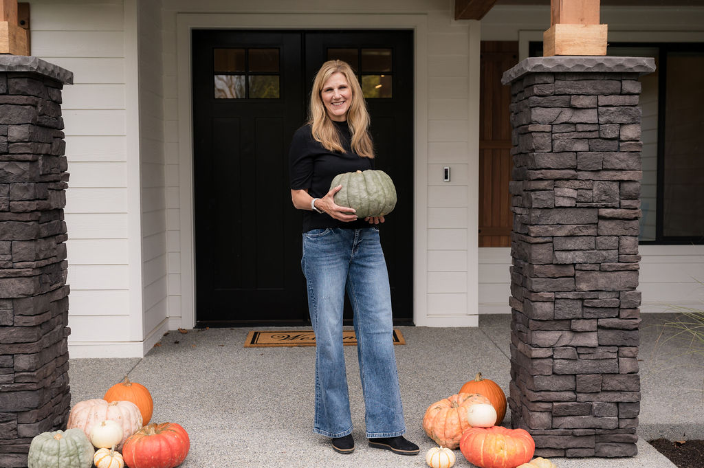 Michelle Yorke standing outside a home holding a pumpkin, decorating for Fall. The home is white with black details and natural stone pillars. 